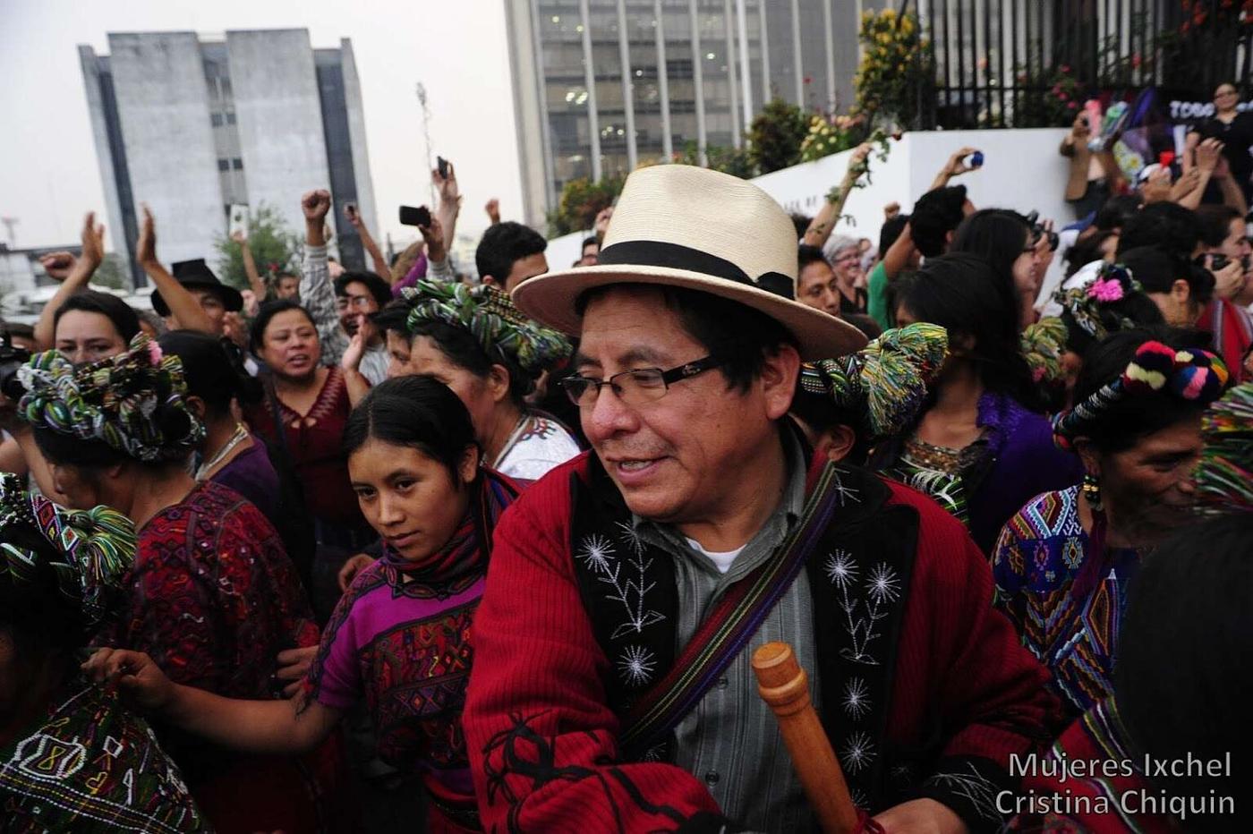 Multitud congregada al aire libre. En primer plano destaca un hombre maya sonriente con sombrero, anteojos y saco tradicional rojo, sosteniendo una vara de autoridad.