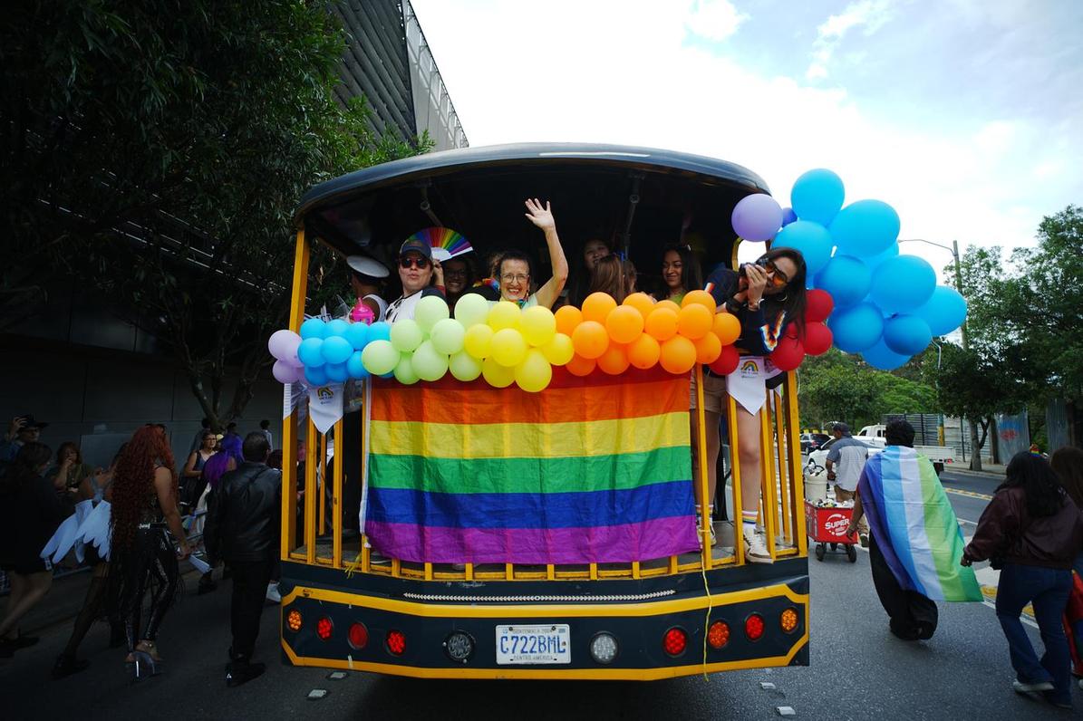 Personas saludando desde la parte de atrás de un furgón. Llevan banderas y globos con los colores del arcoiris.