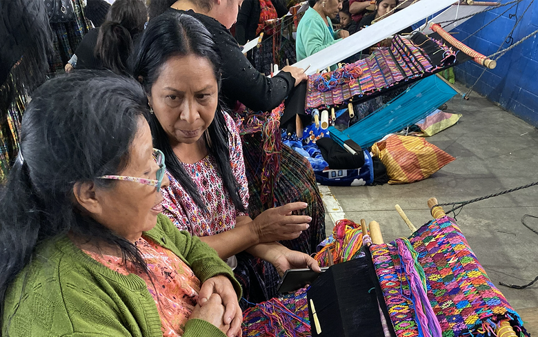 Dos tejedoras indígenas; una de ellas trabajando en un telar de cintura con hilos multicolores mientras la otra mujer la observa de cerca.